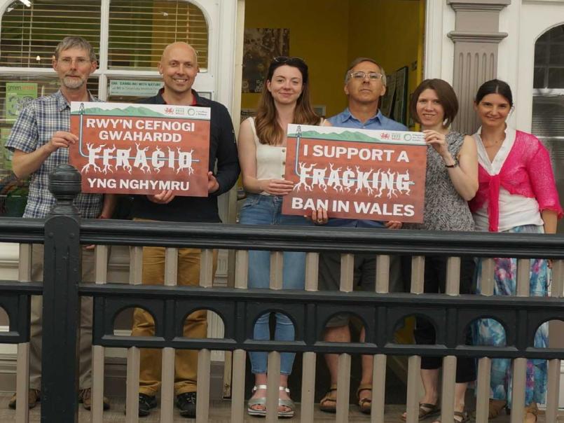 6 people standing side by side in a balcony holding up 'I suppose a fracking ban in Wales' placards